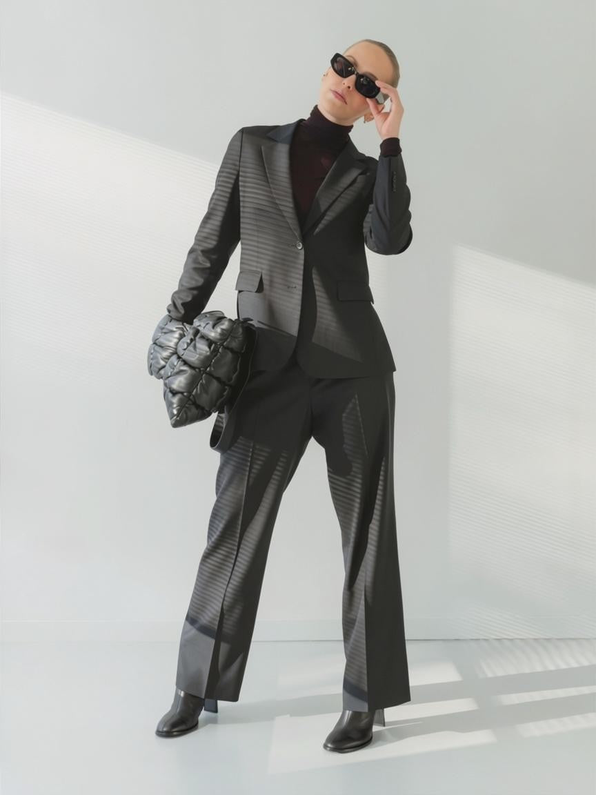 A model wearing a charcoal textured fine twill suit with a black turtleneck, black sunglasses, and holding a black quilted clutch bag against a minimalist white studio background.