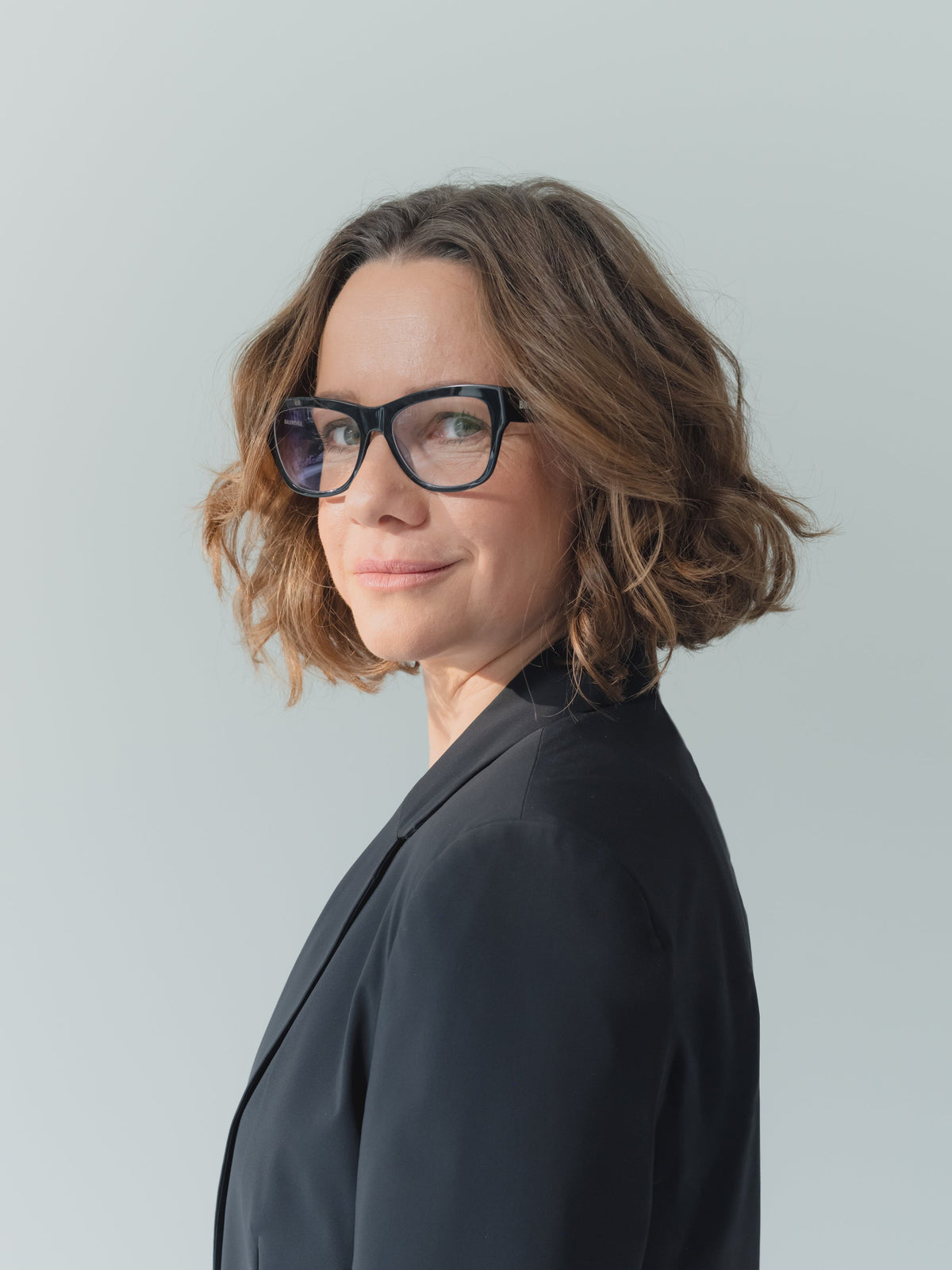 Antje Bleek with short wavy brown hair and black-rimmed eyeglasses, wearing a dark blazer, shown in a side-profile portrait against a light gray background.